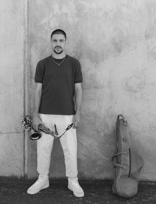 Joshua Moshe black and white photo with wall background. Joshua is holding an alto saxophone